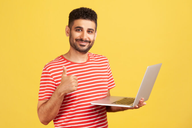 A man in a red striped t-shirt stands against a yellow background whilst holding a laptop and giving a thumbs up