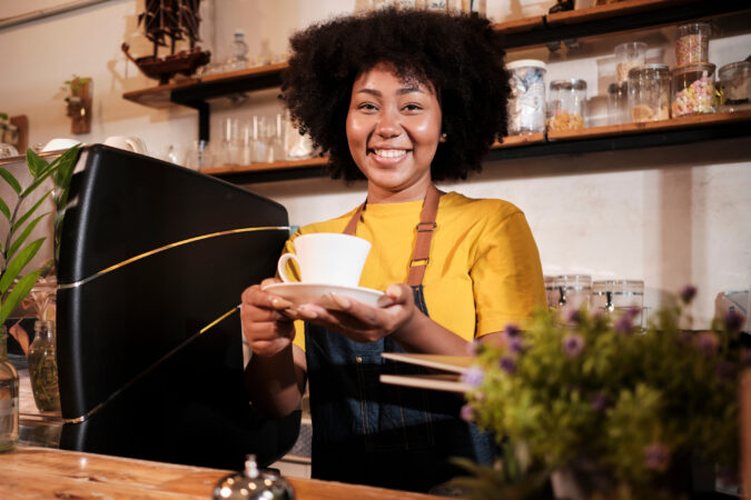 A barista dressed in yellow presents a mug of coffee in her hands